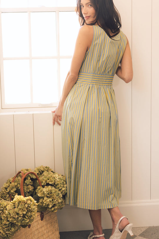 A woman wearing the Ivy City Co Glen Dress in Green Stripe—a sleeveless, cotton, ankle-length dress with yellow and blue stripes—stands by a window, glancing over her shoulder. On the floor beside her is a straw basket filled with green hydrangeas.