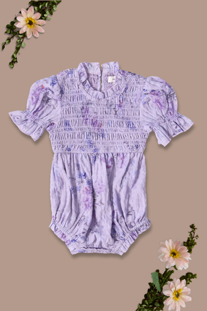 The Ivy City Co Baby Flynn Romper in Lavender Aquarelle Fleur features a smocked bodice, short ruffled sleeves, and a lavender floral print, displayed on a taupe background with pink watercolor flowers and green leaves in the corners.