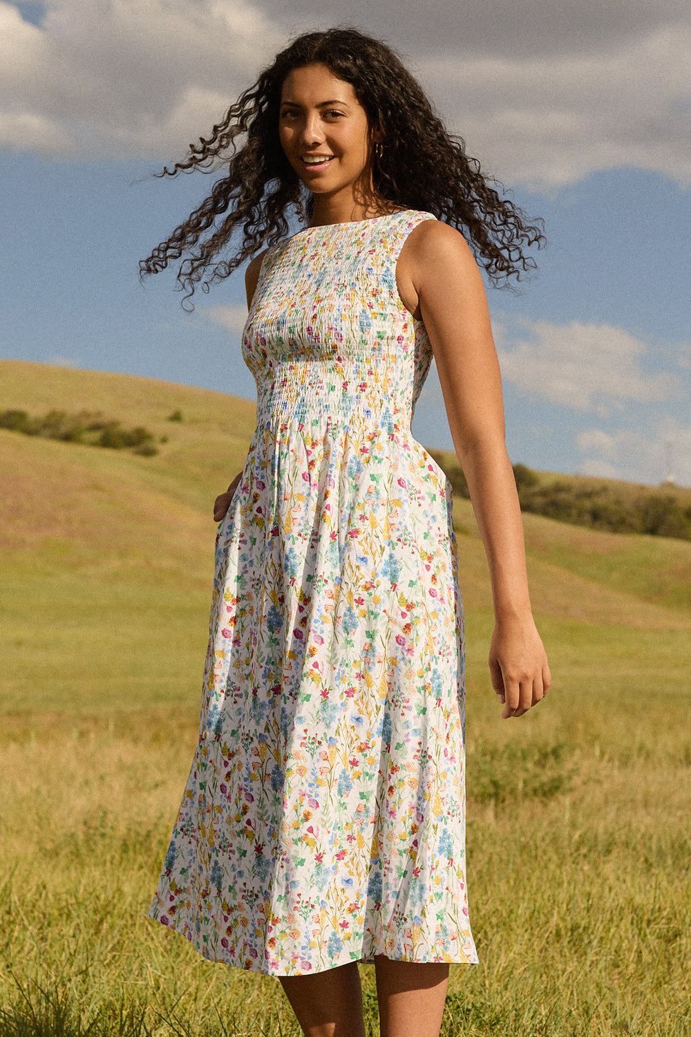 A woman with curly hair stands in a grassy field under a partly cloudy sky, smiling at the camera while wearing the Ana Dress in Floral Fields by Ivy City Co—a sleeveless, floral-print midi dress featuring a smocked bodice.