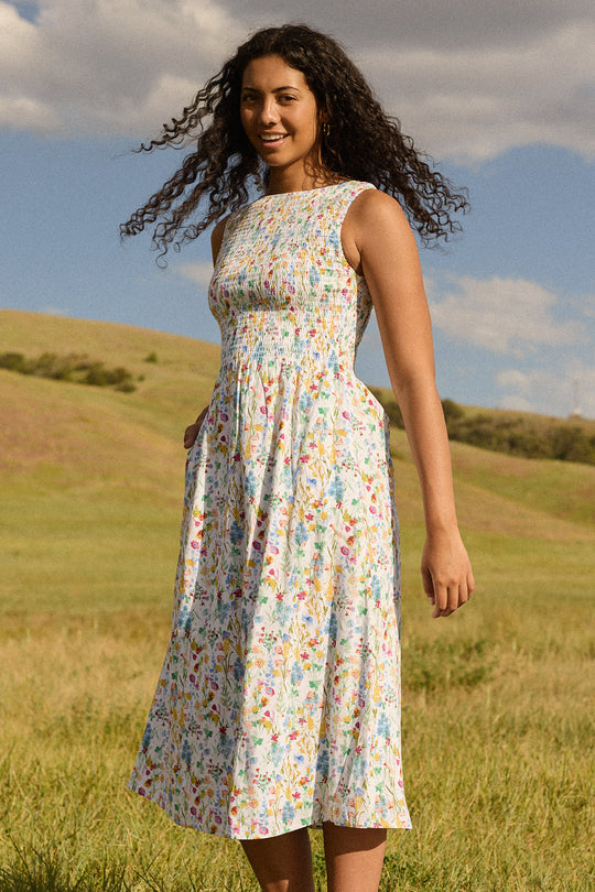A woman with curly hair stands in a grassy field under a partly cloudy sky, smiling at the camera while wearing the Ana Dress in Floral Fields by Ivy City Co—a sleeveless, floral-print midi dress featuring a smocked bodice.
