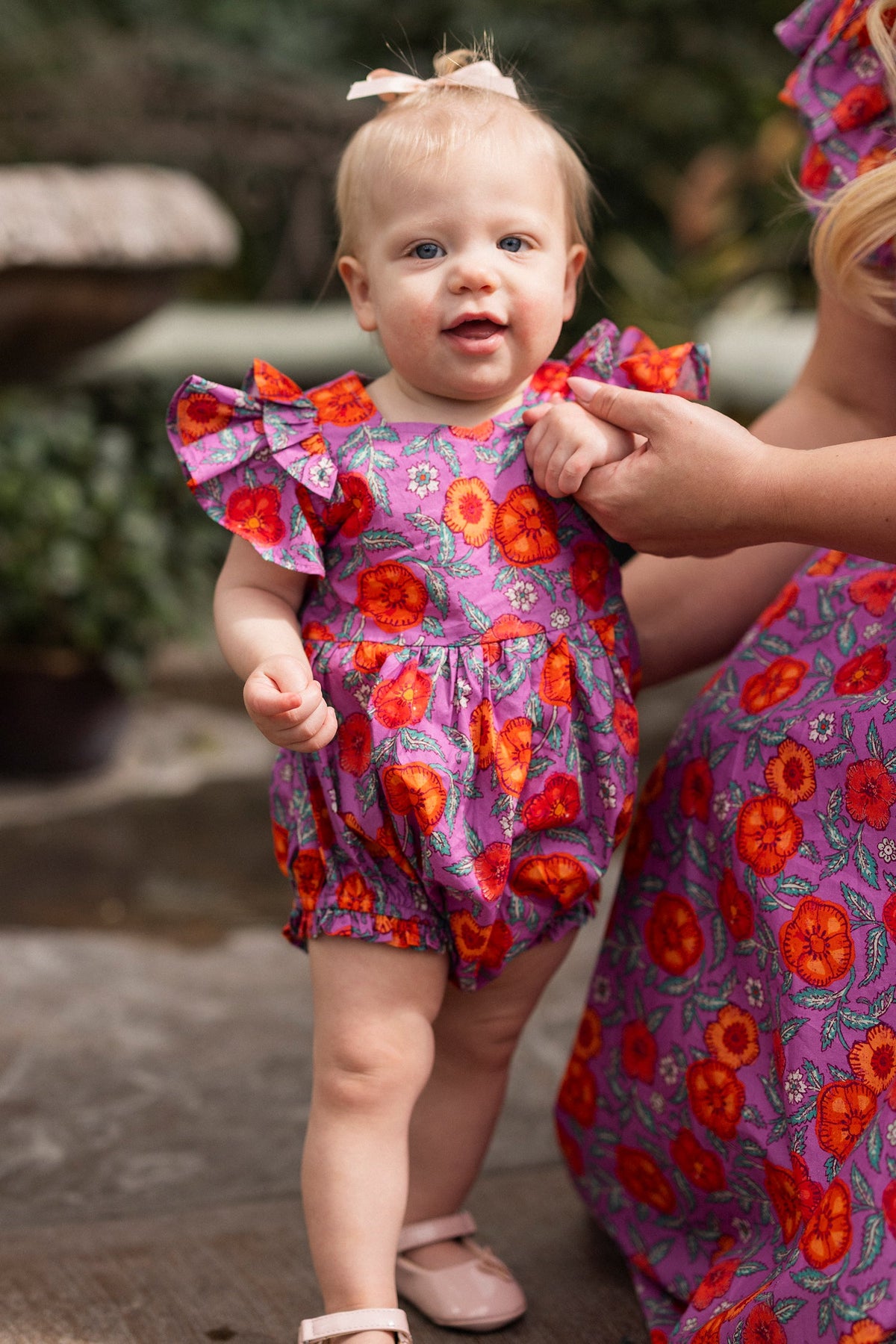 A baby with light hair and a bow stands outdoors near greenery, wearing the soft cotton Baby Hattie Romper in Thistle Flower Poppy Woodblock by Ivy City Co, while holding an adult’s hand. The adult wears a matching floral dress.