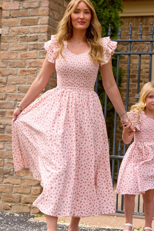 A woman wearing the Ivy City Co Hattie Dress in Madeline Pink, a light pink floral short-sleeve cotton midi dress, holds a girl’s hand. They stand outside near a brick wall and metal fence, both dressed in matching Ivy City Co birthday girl dresses.