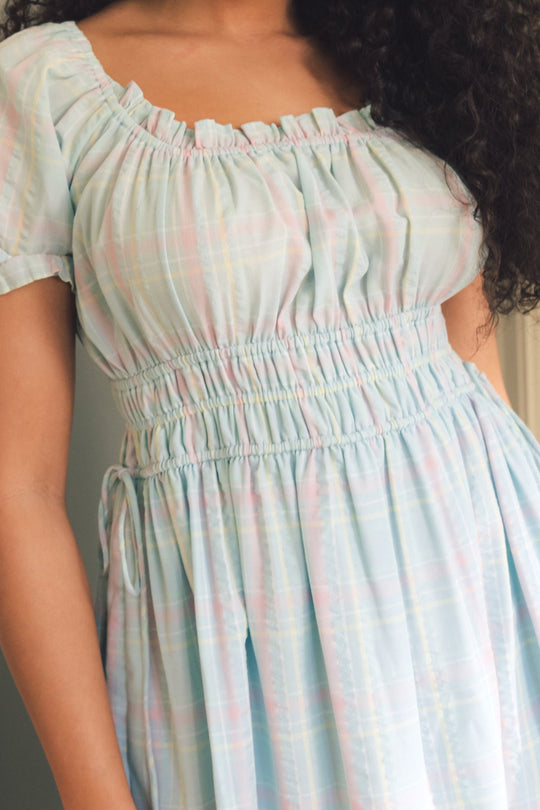 A woman models the Ivy City Co Thalia Dress in Pastel Plaid, showcasing its light blue tones and soft, feminine plaid pattern. The dress features short sleeves, a smocked waist for comfort, a ruffled neckline, and gathered fabric details—perfect for spring. Her curly hair cascades over her shoulders, enhancing the fresh and charming appeal of this stylish piece.