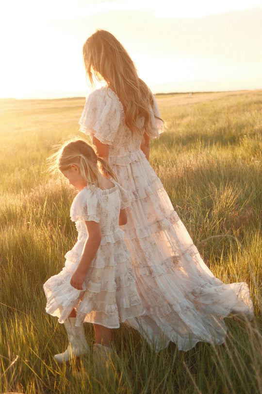 A woman and a young girl in matching white lace dresses—her little one twirling in Ivy City Co’s Mini Winnie Dress in Bright White Wispy Wildflower, the perfect girls’ white floral dress—walk through a sunlit grassy field, sunlight softly illuminating their hair and clothing.