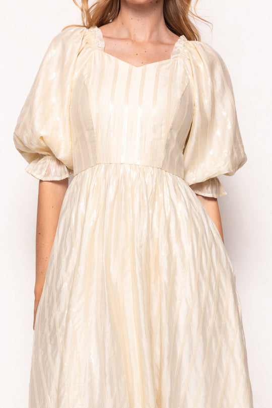 A woman models the Ivy City Co Tindra Dress in Cream Sequin—a cream-colored, puff-sleeve midi dress featuring subtle vertical stripes, a square neckline, and a fitted waist with a soft sheen. Maternity friendly for added comfort; the image is cropped at the elbows and above the chin.