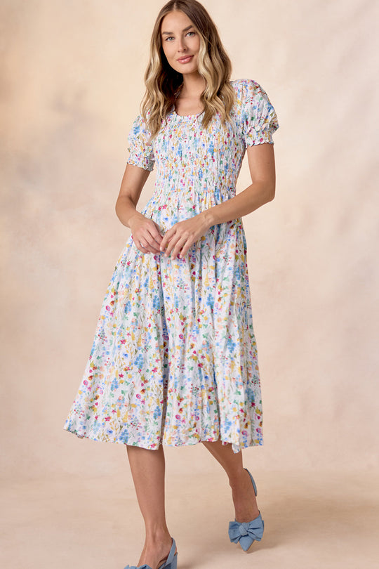 A woman stands against a soft beige background wearing the Amelie Dress in Floral Fields by Ivy City Co—a light blue, floral-patterned, short-sleeve, smocked midi dress—paired with blue high-heeled shoes.