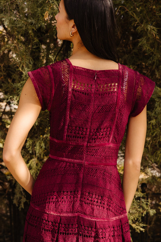 A woman with long dark hair stands outdoors, facing away, wearing the Ivy City Co Amory Dress in Burgundy. The dress features a square neckline, short sleeves, and intricate lace patterns. Green foliage is visible in the background.