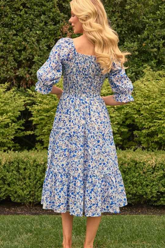 A woman with long blonde hair, seen from behind, stands on grass wearing the Ivy City Co Madeline Dress in Ditsy Cobalt Floral. She is outdoors, surrounded by green bushes and plants.