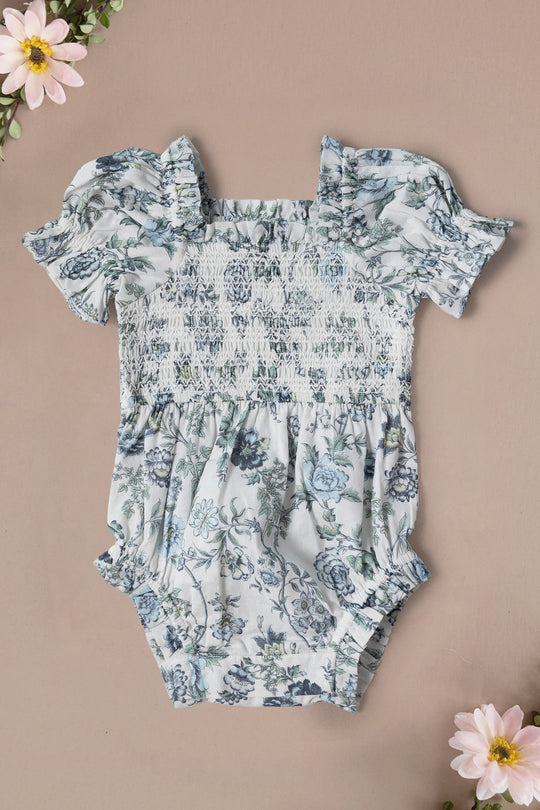 Ivy City Co’s Baby Valencia Romper in Ivory Climbing Vine Bloom features ruffled sleeves, a smocked bodice, and a gathered waist, shown on a beige background accented with pink flowers in the corners.