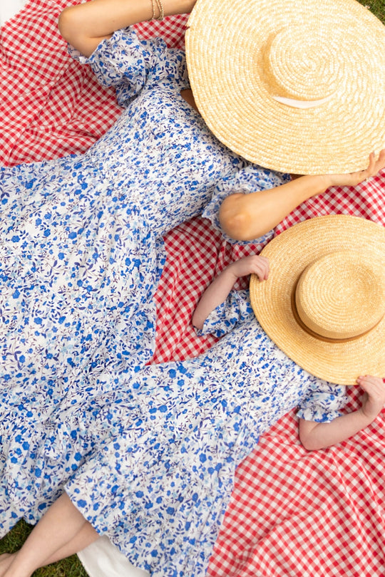 Wearing matching Ivy City Co Madeline Dresses in Ditsy Cobalt Floral, two people lie on a red checkered blanket outdoors, covering their faces with large straw hats as they relax and enjoy each other’s company.