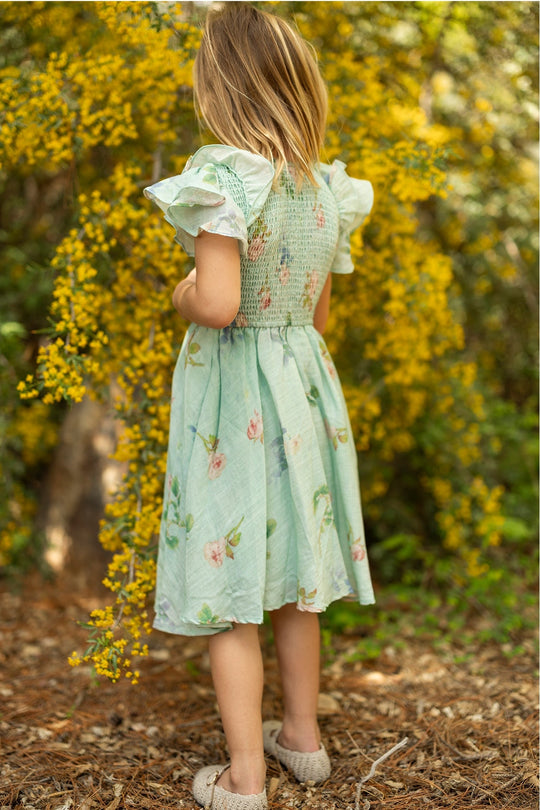 A young girl in the Ivy City Co Mini Hattie Dress in Roselyn Blue stands outside with her back to the camera, surrounded by bright yellow flowers and greenery on a sunny day.