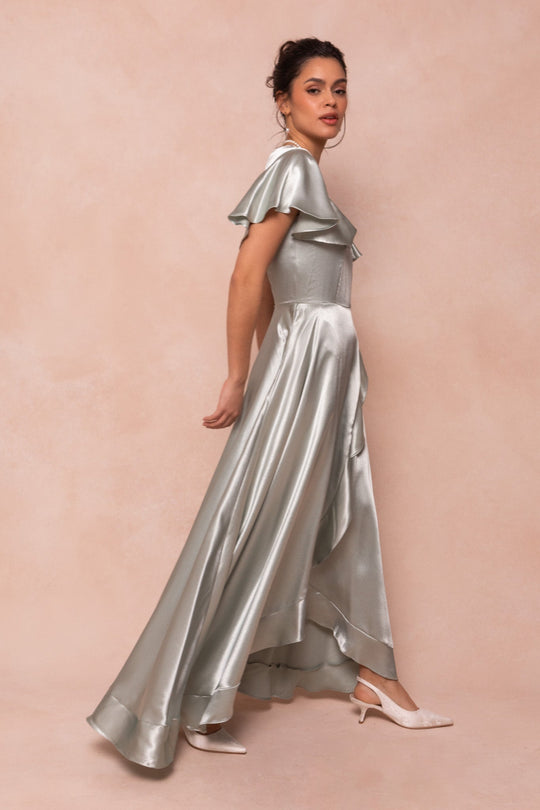 A woman poses sideways against a soft beige background, wearing the Ivy City Co Callie Dress in Sage Satin—an elegant floor-length gown with ruffled short sleeves—paired with white heeled shoes.