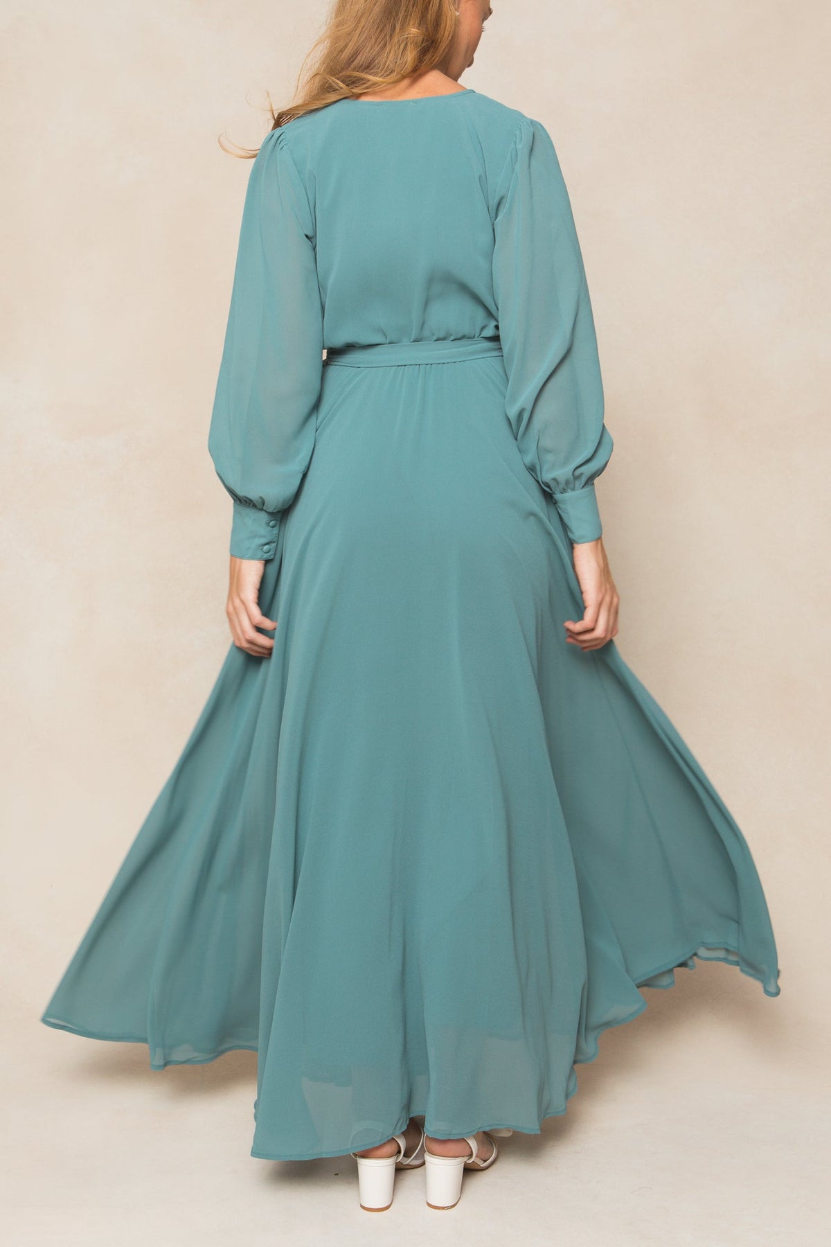 A woman with long hair is seen from behind wearing the Ivy City Co Andie Dress in Seaglass Chiffon, which features long sleeves and a flowing silhouette. She pairs the dress with white heeled sandals.