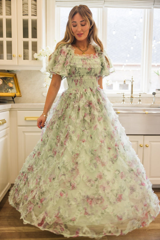 A woman stands in a kitchen, looking down and gently holding the skirt of the Ivy City Co Florentina Dress in Pistachio Aquarelle Fleur. This long chiffon dress features delicate pink floral patterns and ruffle details, with light streaming through large windows behind her. Designed to be maternity friendly, it offers both comfort and elegance for expectant mothers.