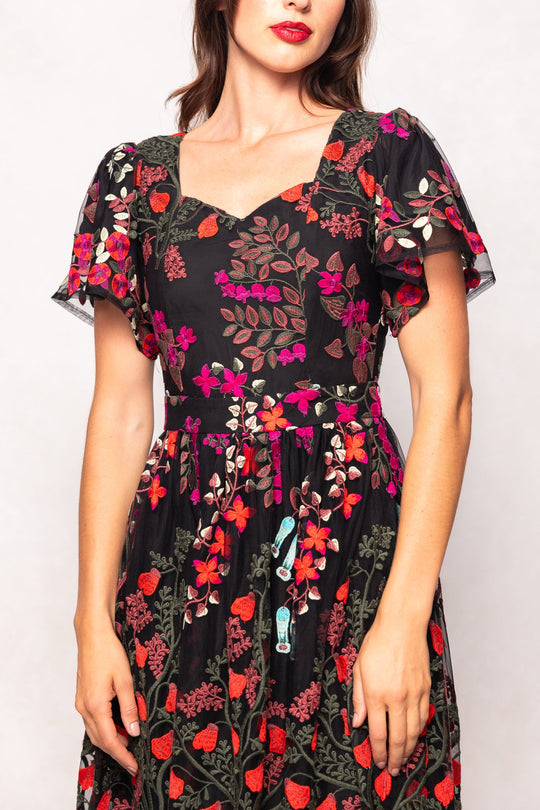 A woman wearing the Ivy City Co Flora Dress in Moonlit Blossom — a black dress adorned with colorful embroidered floral blossoms, featuring short flutter sleeves and a square neckline. She has wavy brown hair and red lipstick, standing against a plain light background.