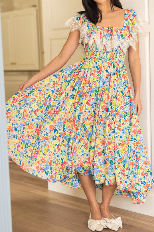 Wearing the Ada Dress in Vibrant Yarrow Print by Ivy City Co—a colorful, floral chiffon dress with delicate lace trim—a woman holds out the skirt indoors on wooden flooring. She pairs it with white bow shoes, and her face remains unseen.
