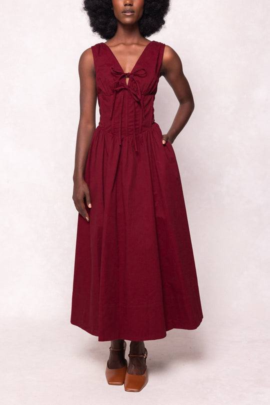 A woman stands against a plain, light background wearing the Kayla Dress in Burgundy by Ivy City Co—a sleeveless midi dress with a v-neckline, bow detail on the chest, fitted bodice, and gathered skirt—paired with light brown heeled shoes.