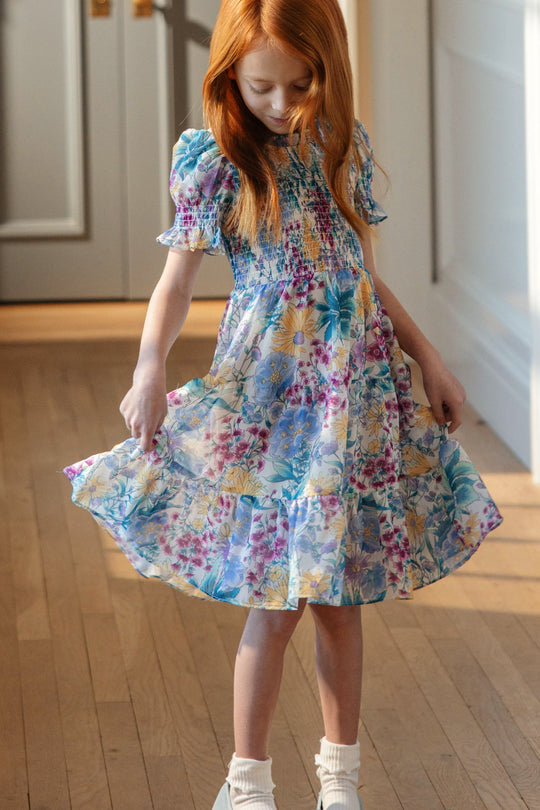 A young girl with long red hair wears the Ivy City Co Mini Cordelia Dress in Pretty Plumeria paired with white socks. Standing indoors on a wooden floor, she looks down while holding out her dress—capturing the joyful essence of spring celebrations and cherished family moments.