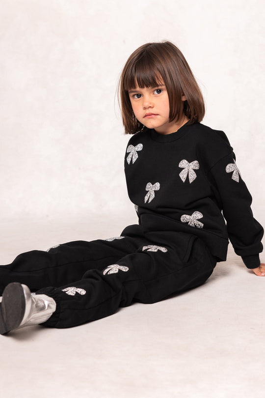 A young child with straight brown hair sits on the floor, wearing a black sweatshirt and matching Ivy City Co Mini Micah Jogger Sweatpants in Black adorned with silver rhinestone bow designs. The background is plain and light-colored.