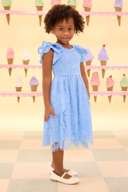 A young girl wearing the Ivy City Co Mini Hattie Dress in Cornflower Blue Lace and white shoes stands smiling in front of a pastel ice cream cone wall display on a checkered floor.