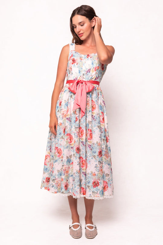 A woman stands against a plain white background, gently touching her hair while wearing the Penny Tank Midi Dress in Multicolor Floral by Ivy City Co—a sleeveless cotton eyelet dress with a pink ribbon at the waist—paired with white mesh shoes.