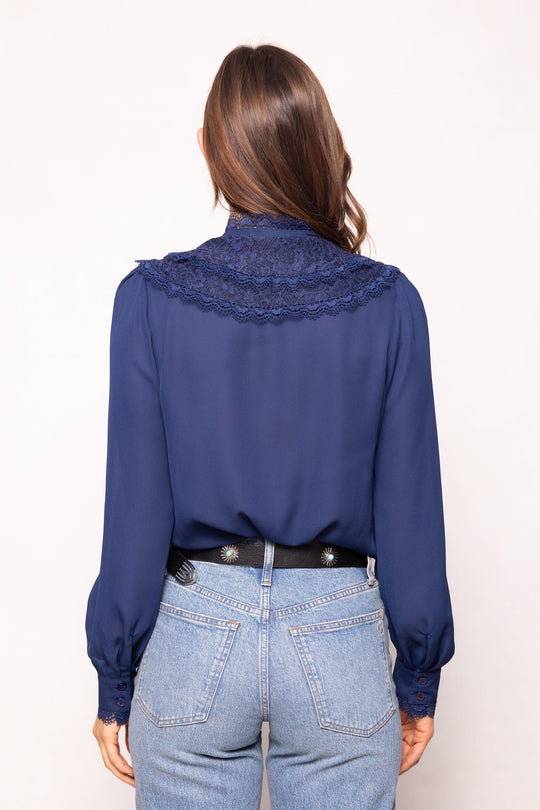 A woman with long brown hair is seen from behind, wearing the elegant Blythe Blouse in Navy by Ivy City Co, which showcases delicate lace detailing on the back and is styled with light blue jeans against a neutral background; this nursing-friendly design beautifully combines style and functionality.