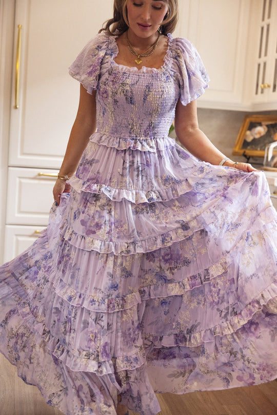 A woman stands indoors in front of white kitchen cabinets and a framed photo, gracefully holding out the skirt of the Ivy City Co Winnie Dress in Lavender Aquarelle Fleur. This flowing, tiered chiffon maxi dress combines timeless style with maternity-friendly comfort.