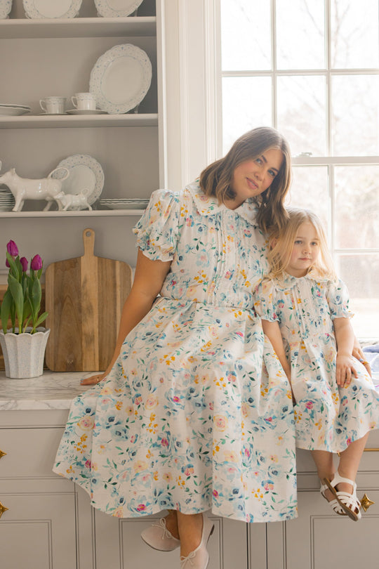 A woman and a young girl, both dressed in the Ivy City Co “Sophia Dress in Blue Bouquet,” sit on a kitchen counter. They are beside a vase of purple tulips, with shelves of white dishes and a large window in the background.