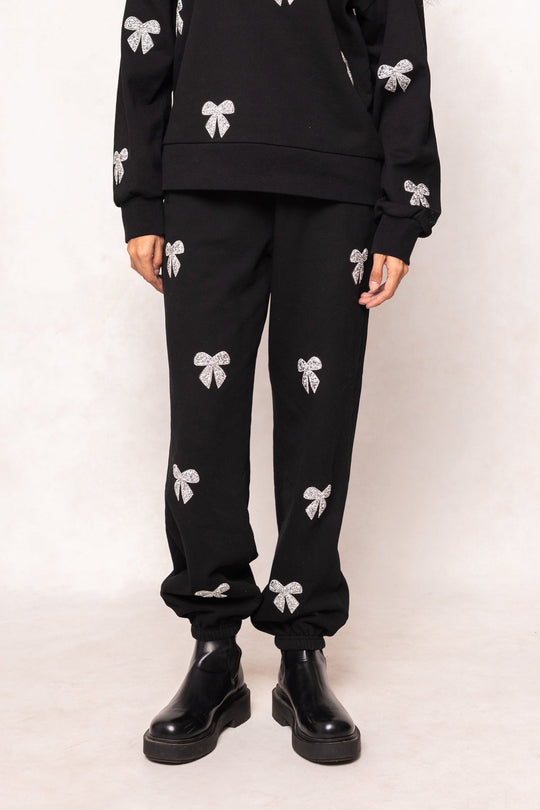 A person wearing Ivy City Co’s Micah Jogger Sweatpants in Black—black joggers embellished with white rhinestone bow designs—paired with black boots, stands against a plain light background. Only the lower half of the body is visible.