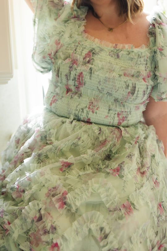 A person wearing the Florentina Dress in Pistachio Aquarelle Fleur by Ivy City Co, featuring delicate pink floral patterns and ruffled details, is seated indoors under soft lighting. This elegant, maternity-friendly dress blends comfort and style, making it ideal for special occasions.