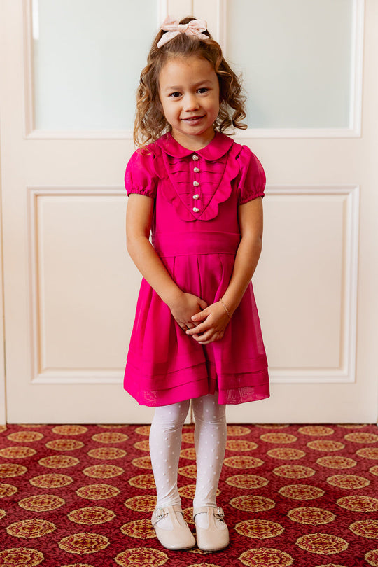 A young girl smiles with her hands clasped in front of her as she stands on a red patterned carpet, wearing the Ivy City Co Mini Cassian Dress in Hot Pink paired with white tights and shoes.