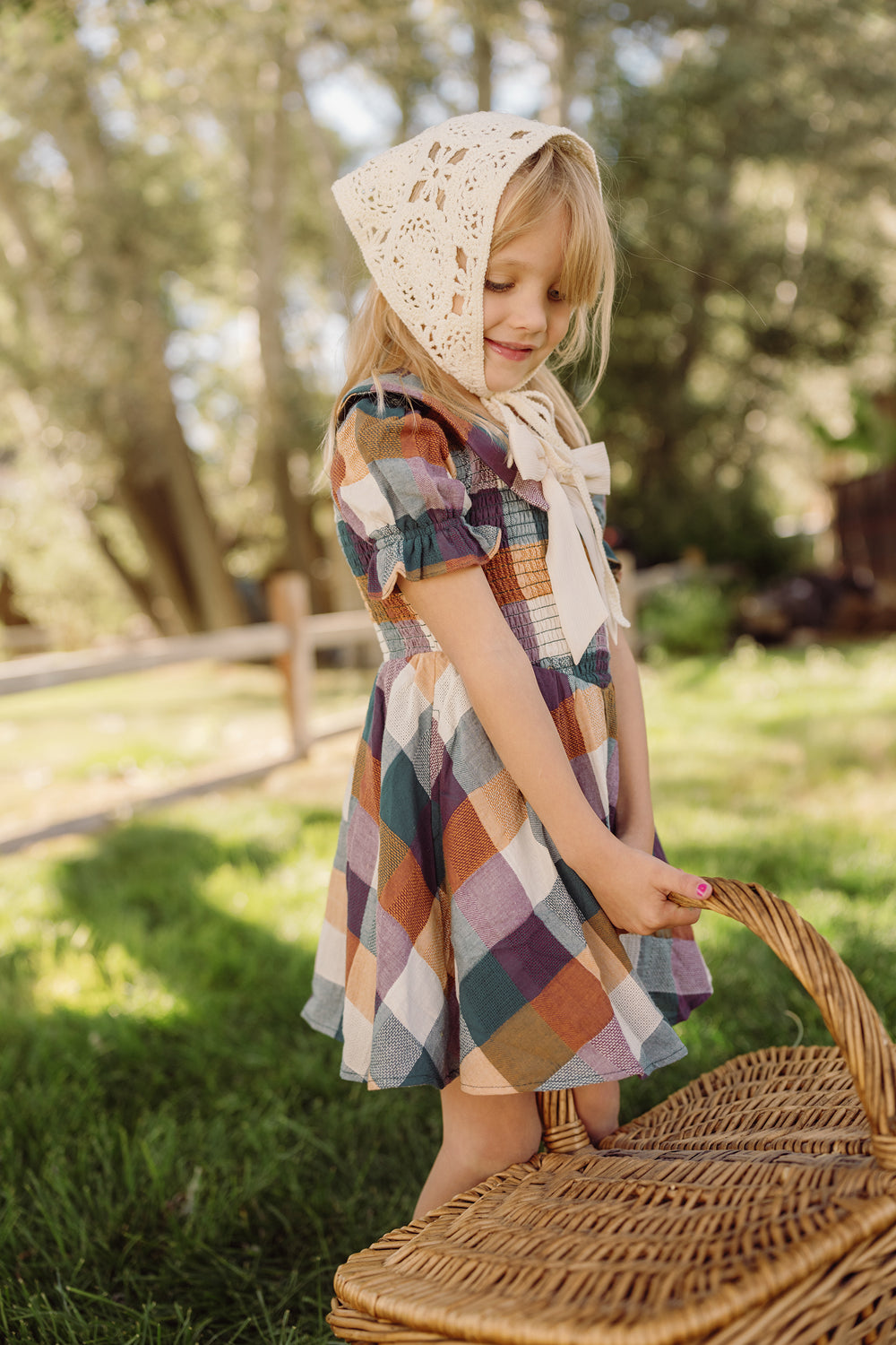 A young girl wearing the Ivy City Co Mini Alice Dress in Maize Gingham, featuring a smocked bodice and puff sleeves, stands on the grass outdoors holding a wicker basket. Sunlight filters through the trees behind her, illuminating her crocheted bonnet.
