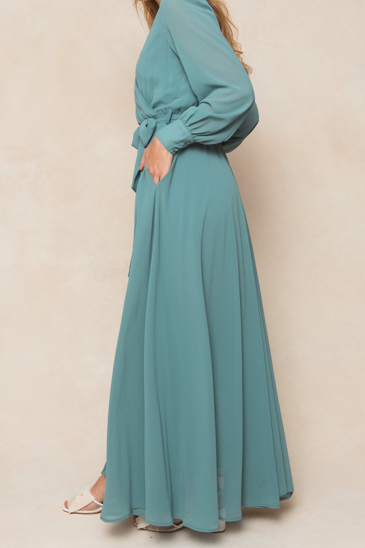 A woman poses sideways with one hand in her pocket, wearing the Ivy City Co Andie Dress in Seaglass Chiffon—a long-sleeved, maxi-length dress with a waist tie. She pairs it with white open-toed shoes against a neutral background.