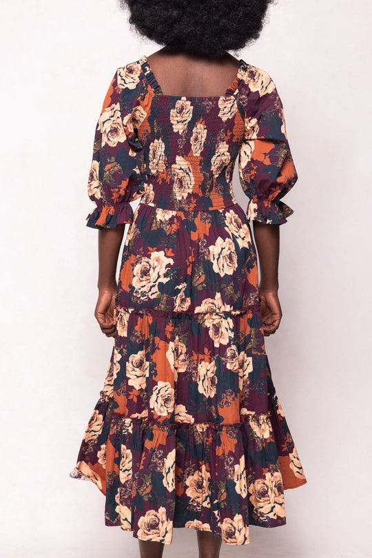 A woman with curly hair wears the Ivy City Co Madeline Dress in Equinox Rose, a floral midi dress featuring puffed sleeves and a square neckline, shown from the back against a plain light background.
