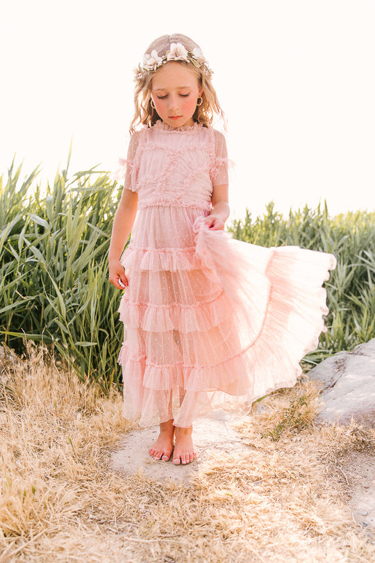 A young girl stands barefoot on grass, gently holding her Ivy City Co Mini Whimsical Maxi Dress in Blush, featuring pink ruffles and flutter sleeves, with a flower crown atop her head and tall green plants in the background.