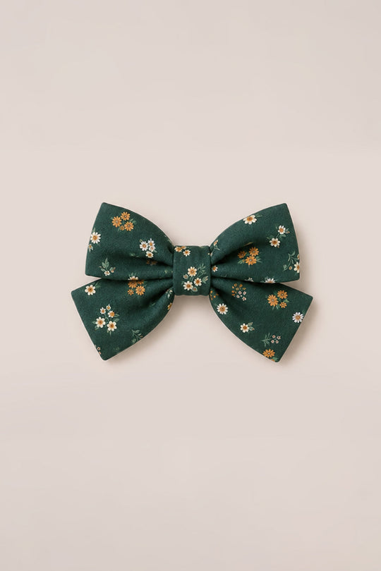 The Hair Bows in Evergreen Lovelorn Ditsy by Ivy City Co feature a dark green cotton fabric decorated with a delightful pattern of small white, yellow, and orange flowers, shown against a light beige background.
