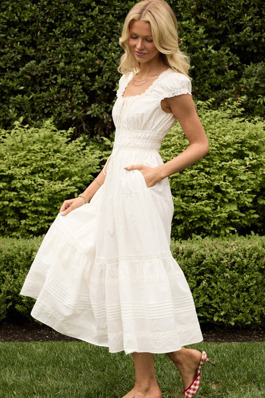 A woman with long blonde hair stands on grass in front of green bushes, wearing the Ivy City Co Palermo Dress in White—a white, short-sleeve, ankle-length flowy dress that she holds at the skirt—and red gingham shoes. The Palermo Dress is both maternity friendly and nursing friendly, providing comfort and convenience for new or expecting mothers.