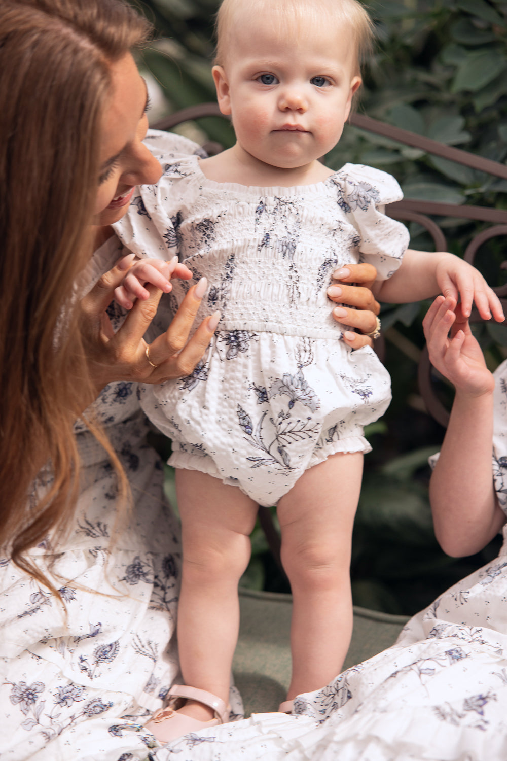 A baby stands while an adult gently holds their hands. Both wear matching light blue, floral-patterned outfits from Ivy City Co—the baby in the Baby Roselyn Romper in Blue Enchanted Floral. Green foliage is visible in the background.