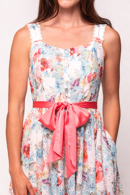 A woman wearing the Ivy City Co Penny Tank Midi Dress in Multicolor Floral, featuring a sleeveless cotton eyelet design with white buttons and a wide pink sash tied in a bow at the waist, stands against a plain background.