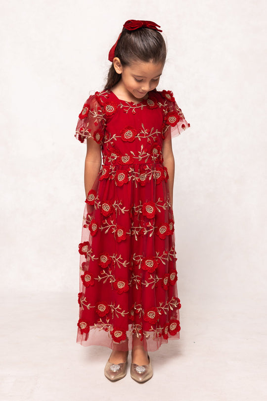 A young girl with brown hair and a red bow looks down, wearing the Mini Athena Dress in Red by Ivy City Co—a girls’ red embroidered maxi dress with short sleeves—paired with silver shoes, and standing against a plain light background. This charming holiday piece is perfect for any special occasion and beautifully embodies the spirit of mommy and me dress styles.