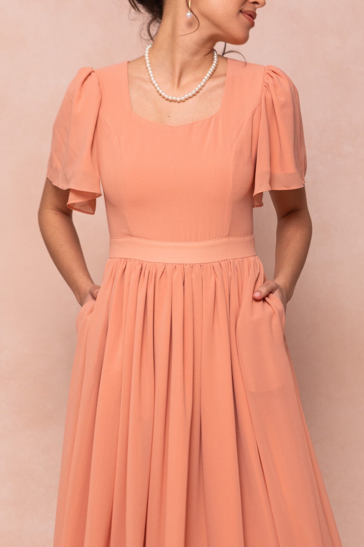 Woman wearing the Abbie Dress in Apricot Crush by Ivy City Co, featuring short flutter sleeves and pockets; styled with a pearl necklace and hoop earrings, she stands sideways against a soft beige background.
