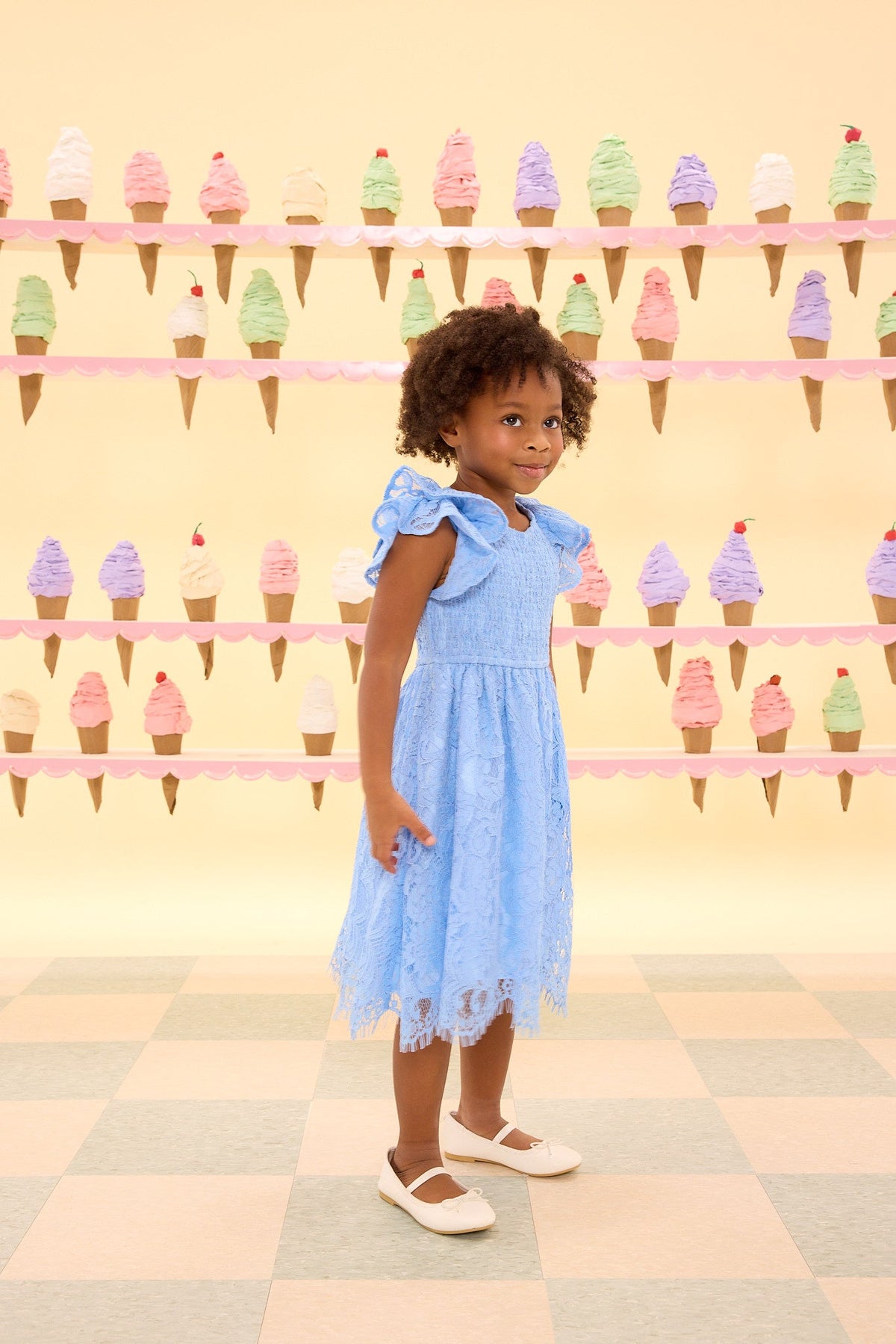 A young girl wearing the Ivy City Co Mini Hattie Dress in Cornflower Blue Lace and white shoes stands in front of a pastel-colored wall decorated with ice cream cone illustrations.