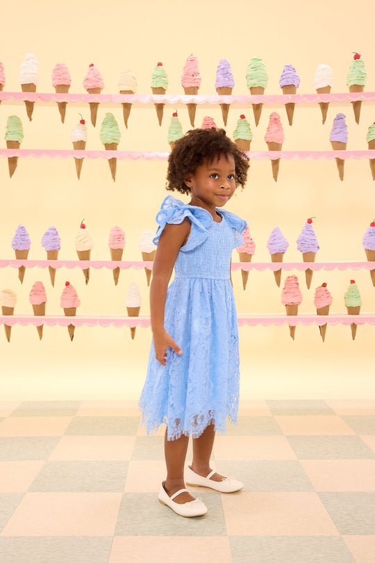 A young girl wearing the Ivy City Co Mini Hattie Dress in Cornflower Blue Lace and white shoes stands in front of a pastel-colored wall decorated with ice cream cone illustrations.