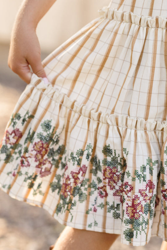 A child holds the hem of the Mini Madeline Dress in Embroidered Cider Grid by Ivy City Co. The skirt is adorned with ruffles and intricate pink and green floral embroidery near the bottom, while the smocked bodice and puff sleeves lend a charming touch to this delightful cream dress featuring tan grid stripes.