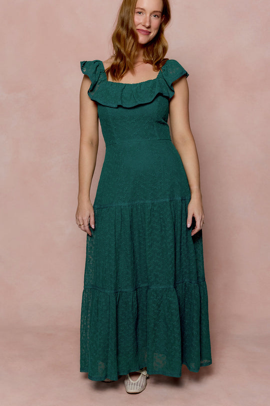 A woman with long brown hair and light skin stands against a pink backdrop, smiling gently as she wears the Ivy City Co Alaina Dress in Deep Sea Lace—a sleeveless, ankle-length dress featuring delicate teal lace, a ruffled neckline, and a tiered skirt. She completes her look with woven beige shoes.