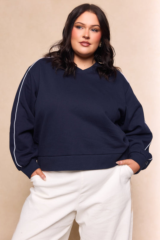 A woman poses confidently in the Ivy City Co Kinsley Sweatshirt in Navy—a cotton fleece style featuring white sleeve stripes—paired with white pants against a neutral beige background.
