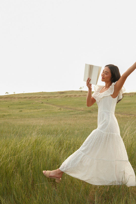 A woman in the Alaina Dress in Ivory by Ivy City Co joyfully walks through a grassy field, holding an open book and smiling with one arm raised, her flowing dress featuring a charming smocked bodice.