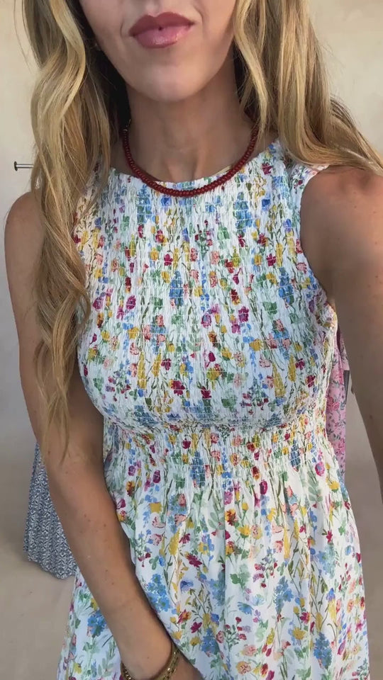 A woman with wavy, light brown hair and a beaded necklace poses indoors against a neutral background, wearing the Ana Dress in Floral Fields by Ivy City Co—a sleeveless, floral-print dress featuring a fitted smocked bodice and loosely draped skirt that’s both maternity friendly and stylish.