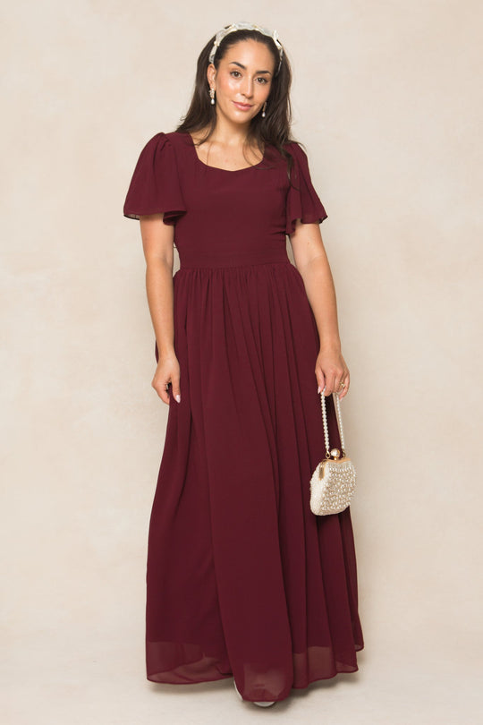 A woman stands against a neutral background wearing the Abbie Dress in Wine Chiffon by Ivy City Co—a burgundy, floor-length gown with short puff sleeves. She holds a beaded purse, her long hair styled with a headband.