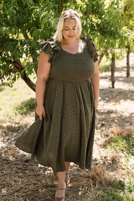 A woman stands outdoors on a sunny day, surrounded by trees and grass, wearing the Ivy City Co Hattie Dress in Woodland Floral. The dress features a smocked bodice and she looks down with a gentle smile. This charming style is both flattering and maternity friendly.